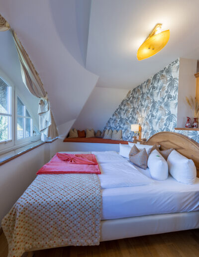 Juniorsuite, vorn: Doppelbett, dahinter gemütliche Sitzecke mit vielen Kissen, links: großes Rundbogenfenster mit Blick in die Natur