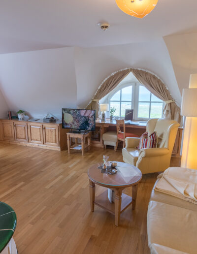 Juniorsuite-Deluxe mit Dachschrägen, Blick in Wohnbereich, rechts: helle Sitzecke, mitte: Rundbogenfenster mit Blick auf Ostsee, links: Kachelkamin