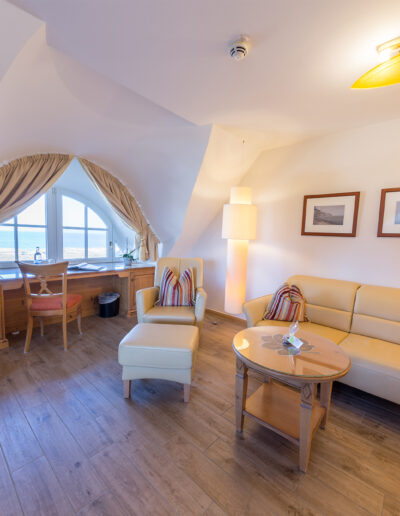 Juniorsuite-Deluxe, Wohnbereich, helle Sitzecke mit Stehlampen und Schreibtisch vor Rundbogenfenster mit Blick auf Ostsee