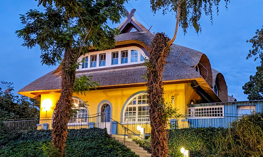 bergfalke-2026 Frontalansicht des gelben Gästehauses mit Reetdach, geschwungenen Fenstern und blauem Fensterrahmen. Das Gebäude ist über eine Treppe mit Geländer erreichbar, im Vordergrund viel Grün.