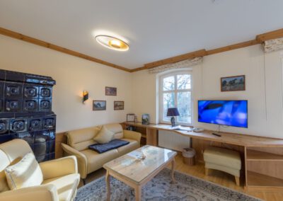 Wohnbereich mit hellem Ledersofa und -sessel, hellem Holztisch und Flachbildfernseher an der Wand; großer Kachelofen am Rand des Raums