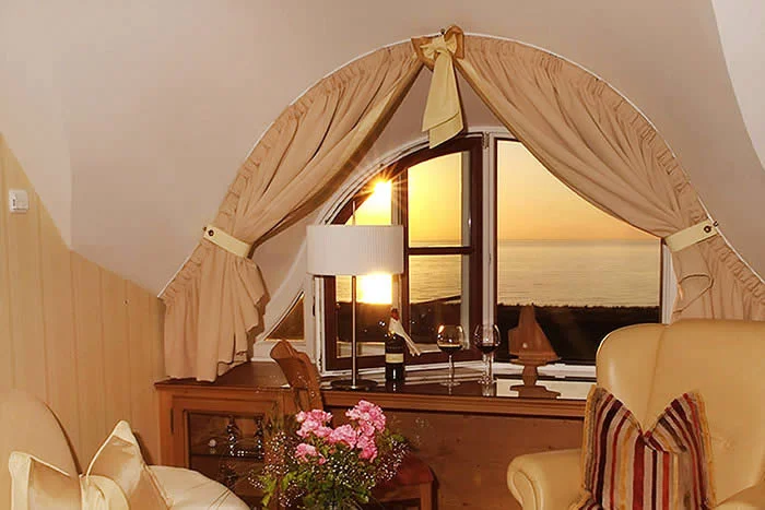 Gemütlich eingerichtete Suite mit gelben Ledersesseln und Blick durch ein Bogenfenster auf das Meer bei Sonnenuntergang.